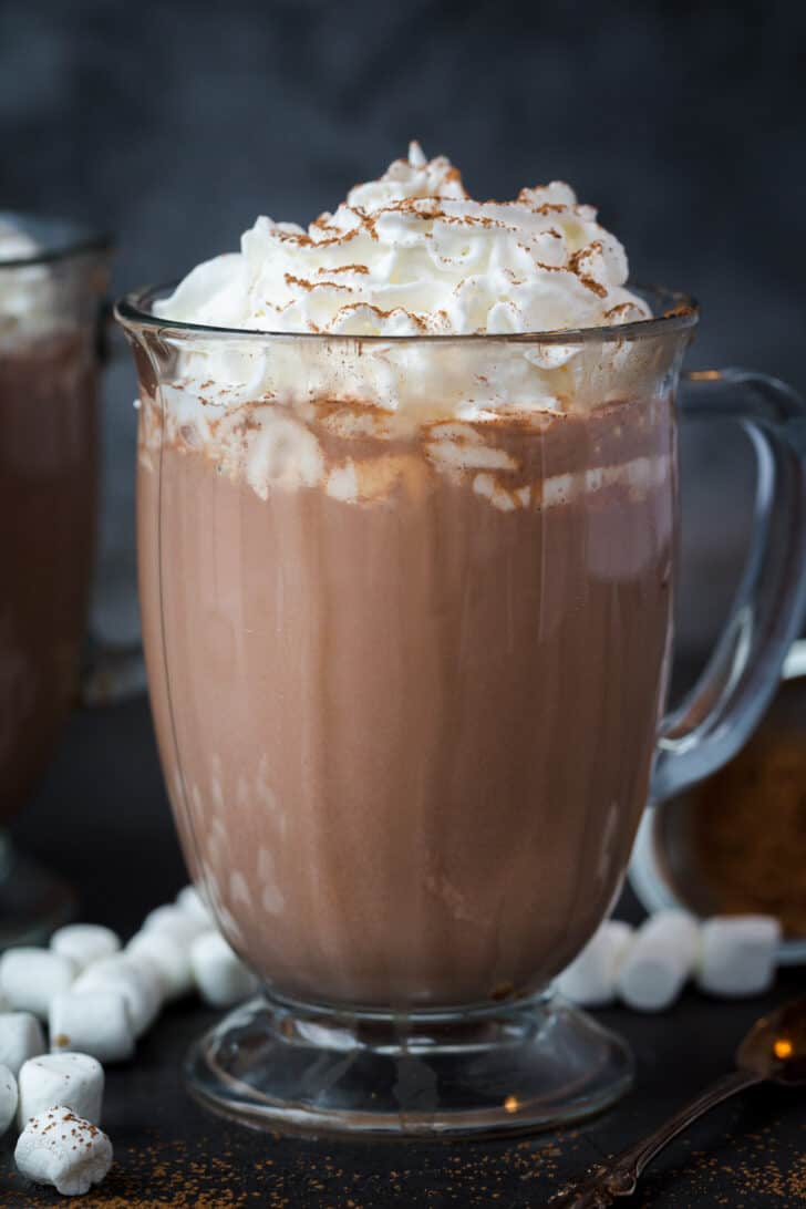 A close-up of hot chocolate in a glass mug with whipped cream and cocoa powder.