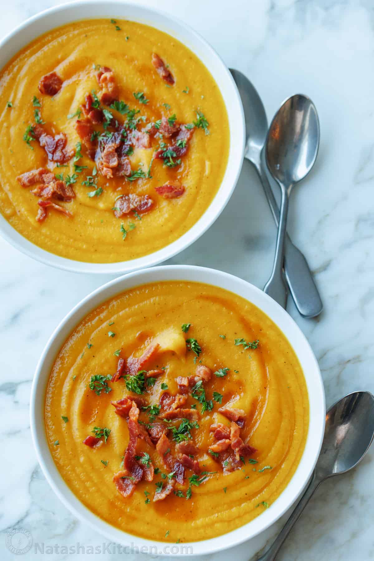 Overhead view of two bowls of butternut squash soup garnished with bacon bits and chopped fresh parsley.