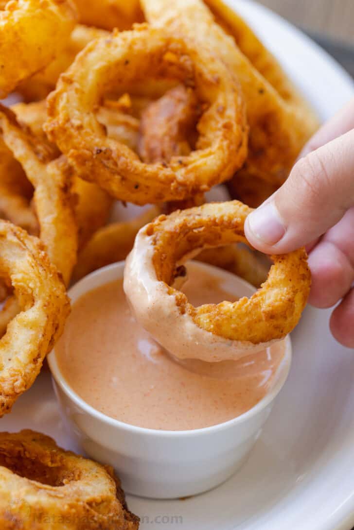 Hand dipping onion ring into small bowl of sauce