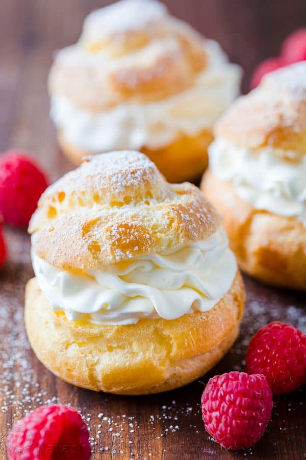 Cream Puffs filled with cream on a cutting board, dusted with powdered sugar, and garnished with raspberries