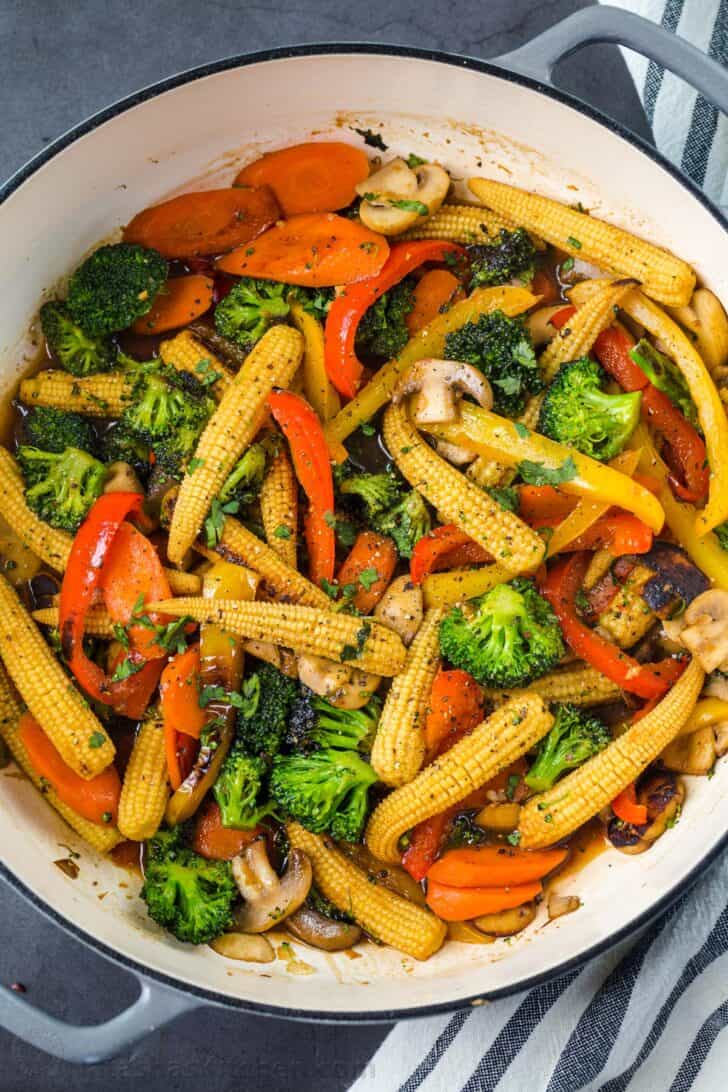 Vegetable stir fry in a skillet topped with greens and a towel on the side of the skillet.