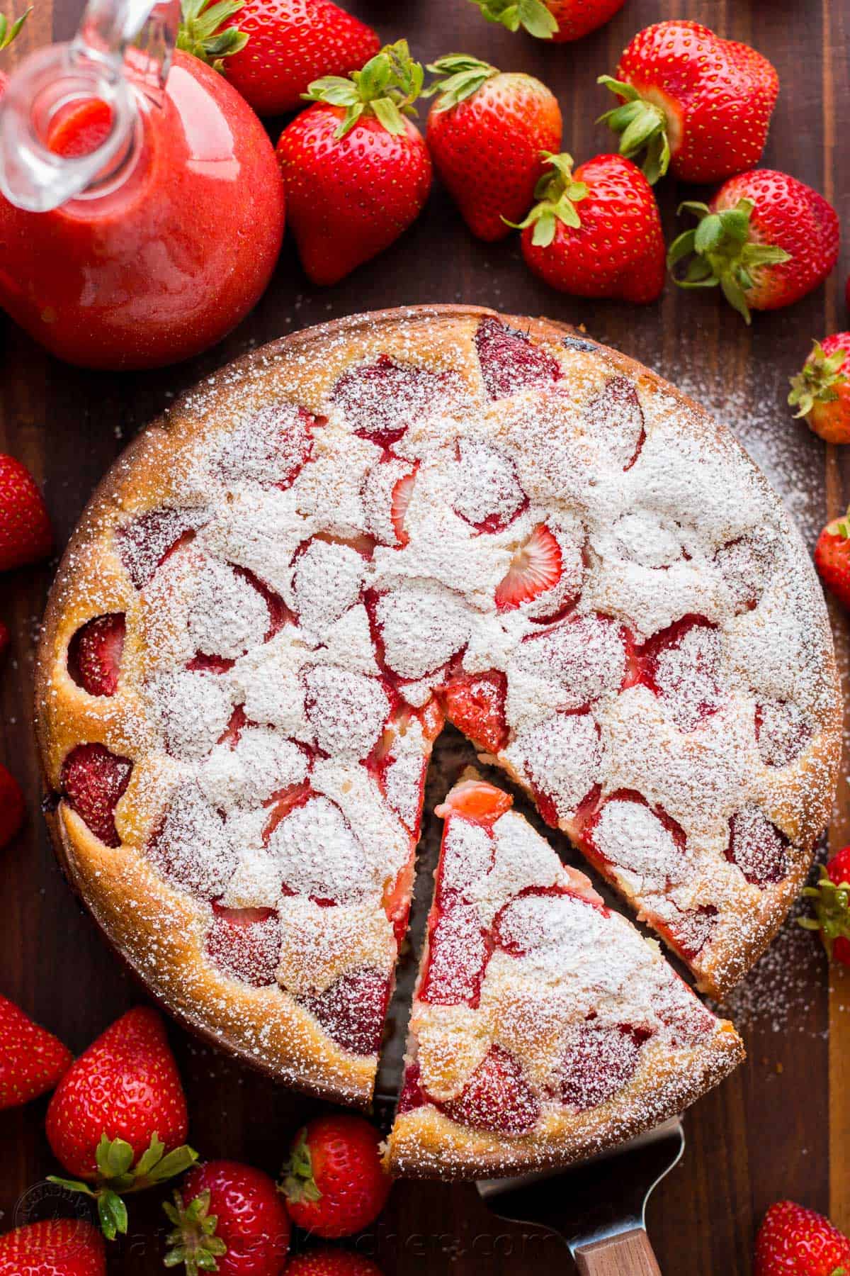 Gâteau aux fraises facile saupoudré de sucre glace avec une sauce aux fraises dans un pichet sur le côté et entouré de fraises fraîches