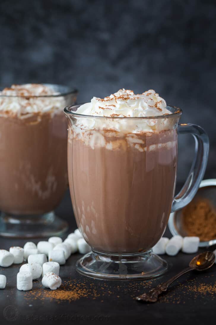 A cup of hot chocolate topped with whipped cream and cocoa powder next to another cup and marshmallows.
