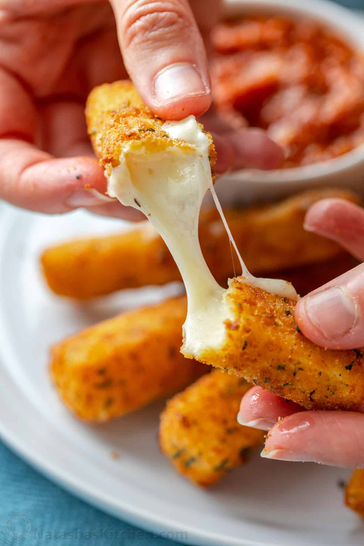 Two hands pulling apart a mozzarella stick with a plate of cheese sticks in the background.