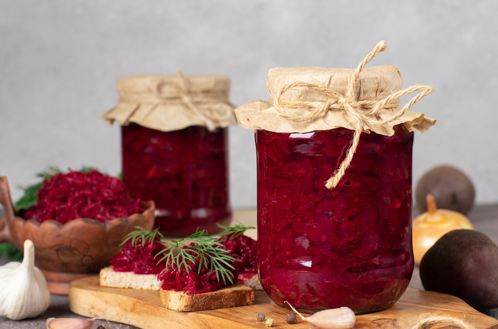 Homemade spicy caviar from beets, carrots, onions and garlic in two jars and bowl on light gray background